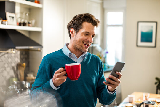 Smiling man texting on smartphone while drinking coffee in home kitchen