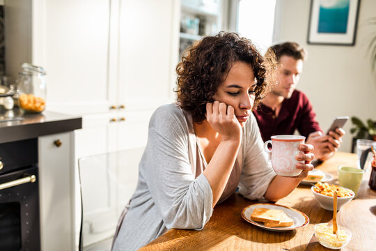 Sad woman at breakfast while partner uses phone in home kitchen