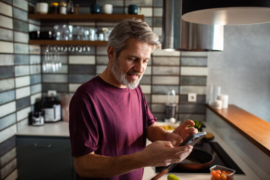 Middle-aged man using smartphone while cooking in modern kitchen
