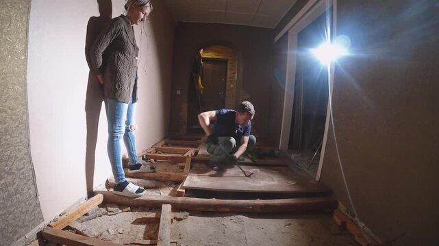 Worker Clearing Debris In Hallway. Caucasian Man Dismantling Floor Joists Under Light. Caucasian Worker Carefully Taking Apart Floor Beams In Confined Corridor Space Under Work Light