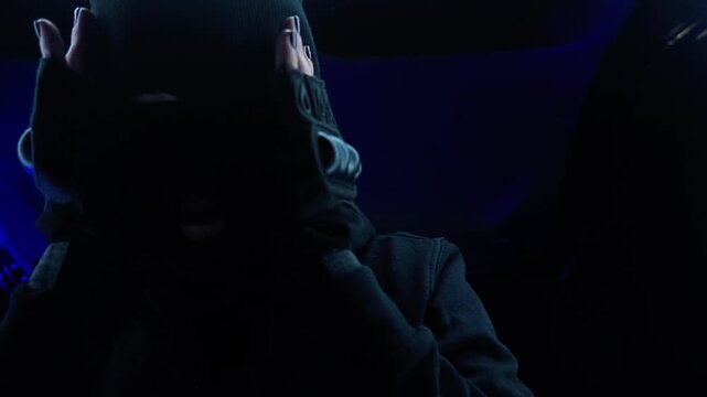 woman bandit robber is going to commit a robbery. puts on a balaclava mask, gloves and a hood in the trunk of a car. night, blue light, atmospheric mysterious shot, preparation