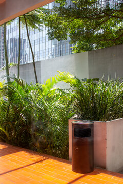 A black iron trash can stands on a red tile path next to a lush tropical garden with an office building background.