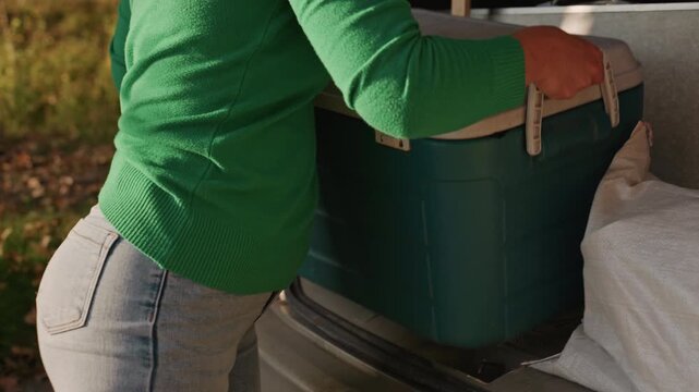 Green cooler being lifted from car trunk for camping setup, woman in green sweater and jeans hauling cooler across leafy campsite, closeup on handles and textured plastic, action shot of preparation