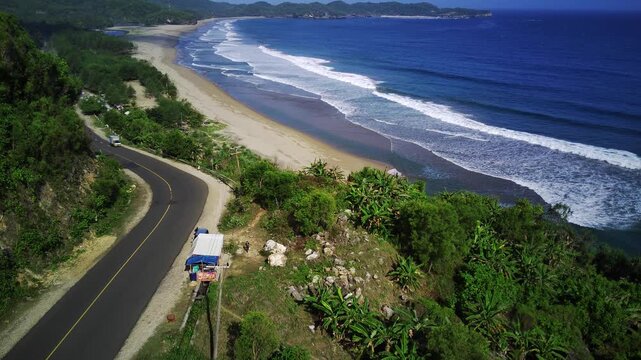 Aerial view of a winding road along a tropical coastline with a white van driving on the asphalt road surrounded by lush greenery and dense foliage overlooking a sandy beach and ocean waves.