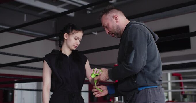 Zoom out footage of a trainer applies protective wraps to a young female boxer's hands, providing protection and support to her wrists and knuckles in preparation for intense boxing training.