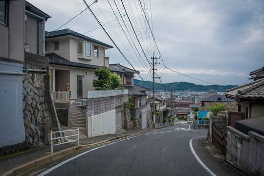 A steep downhill residential street in a Japanese neighborhood looking out toward a distant city harbor and coastal mountains under a cloudy sky.