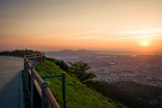 Panoramic sunset view from a mountain observatory overlooking the vast city lights and bay of Kitakyushu as the sun dips below the horizon.