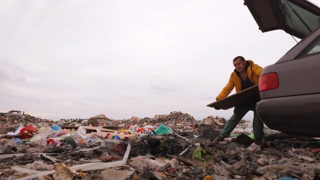 Scavenger in a yellow jacket illegally throwing a wooden board from his car trunk into a large garbage dump, contributing to environmental pollution and contamination with plastic waste