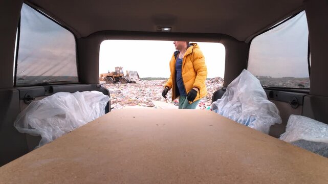 Caucasian Man In Yellow Jacket Lifting Board From Van At Recycling Dump, Straining To Unload Large Sheet, Machinery And Piles Of Waste In Background, Gritty Urban Salvage Scene