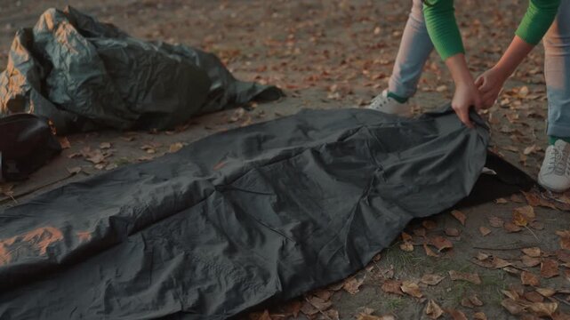 Unfolding tent fabric on leaves, camping camper prepares shelter at dusk with focused hands in jeans and sneaker, smoothing inner liner, checking zipper and pole placement, staking edges and testing