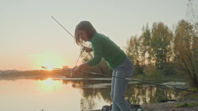 Woman assembling tent poles by lake, attaching connectors and pegs on pebbled shore while securing fabric and cords, concentrated movements in twilight glow, handson gear work, serene treelined
