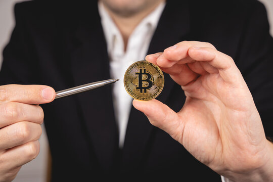 Une main montre une pi&egrave;ce de Bitcoin dor&eacute;e avec un stylo. En arri&egrave;re plan, une personne en costume et chemise blanche.