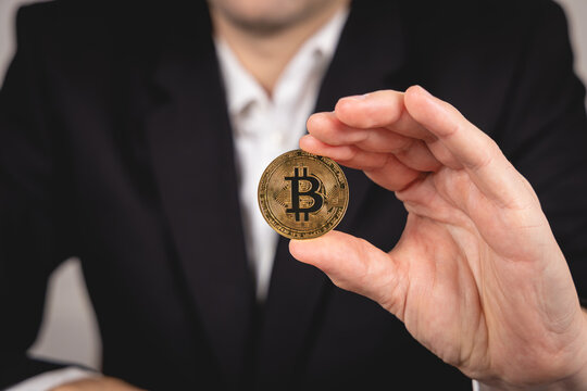 Une main tient une pi&egrave;ce de Bitcoin dor&eacute;e. En arri&egrave;re plan, une personne en costume et chemise blanche.