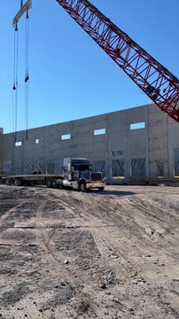 Large truck with flat bed trailer passing by red crane and precast concrete panels warehouse construction site