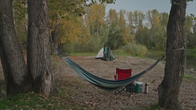 Trees framing hammock campsite autumn dusk, red chair and green tent beyond, camping stove and gear at foreground, cool evening tones, prep for night trek or basecamp rest, tranquil wilderness mood