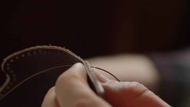 A very detailed close-up shot focusing on the two needles and thick, waxed thread as a craftsman performs a traditional saddle stitch on natural brown leather.