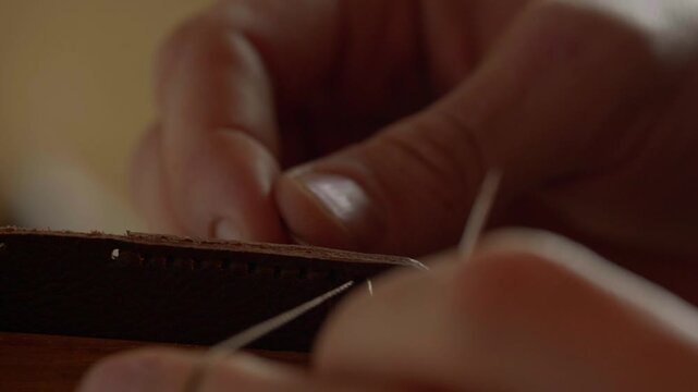A very detailed close-up shot focusing on the two needles and thick, waxed thread as a craftsman performs a traditional saddle stitch on natural brown leather.