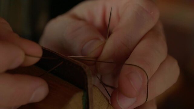 A very detailed close-up shot focusing on the two needles and thick, waxed thread as a craftsman performs a traditional saddle stitch on natural brown leather.