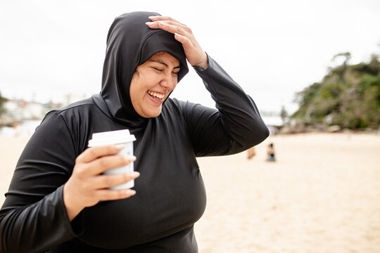 Woman in black burkini laughing while holding a cup of coffee with other hand on her head