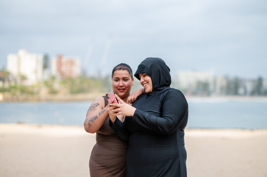 Woman in burkini showing her phone to her friend on the sandy shore at the beach