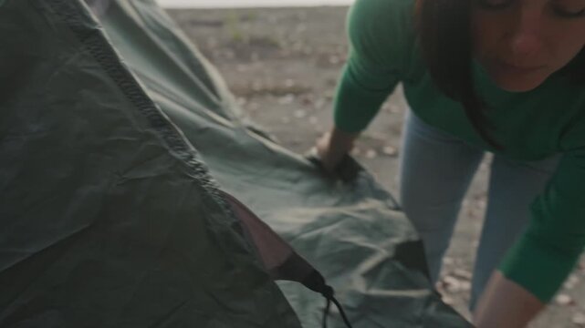 Lakeside sunset camping with caucasian woman unfolding green tent at pebbled shore, fastening zipper and anchoring stake, wearing green sweater and jeans, preparing solo campsite amid calm water
