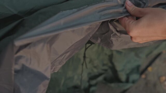 Hands zipping tent fabric closeup dusk, camper tightening slider along nylon seam, instructor style demonstration, focus on fingers, texture of green canvas, careful repair and secure fastening