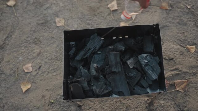Preparing coals for outdoor cooking. Setting up charcoal fire on soil for cooking during camping trip. Assembling and lighting campfire using charcoal and leaves for outdoor meal preparation