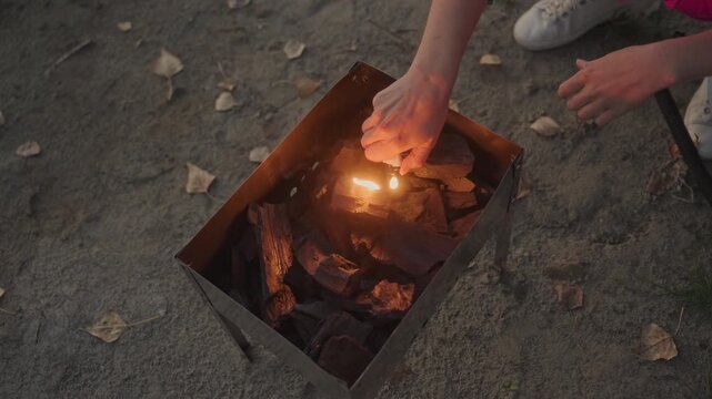 Hands lighting charcoal in portable brazier closeup white camper uses lighter to ignite coals on sandy ground autumn leaves scattered focused closeup stepbystep ignition warm orange flame emerges