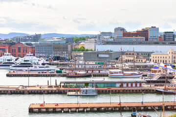 Obraz premium Bustling Oslo Harbour in Norway, featuring traditional sailing ships and modern ferries docked at Aker Brygge. The skyline showcases a blend of historic architecture and contemporary buildings