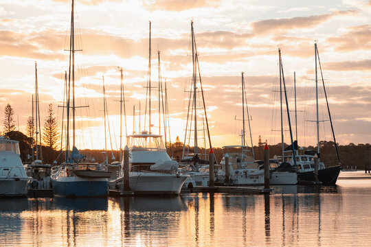 Sailboats and yachts docked at a marina during a golden sunset with reflections on the water