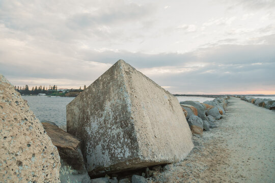 Concrete breakwater blocks line a sandy path leading towards the ocean under a cloudy sky