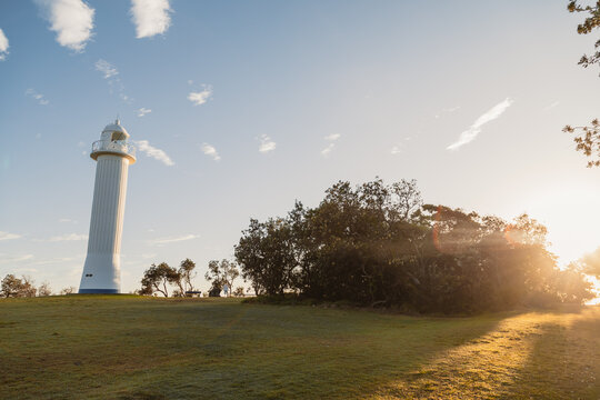 White lighthouse standing tall on a grassy hill at sunrise with sunbeams breaking through trees