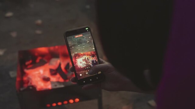 Female camper photographing lake reflection on phone, closeup of smartphone screen showing mirrored sunset over calm water while embers glow in foreground, hand framing shot with grill and coals out