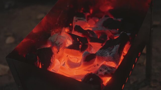 Glowing charcoal coals in camping grill, ember glow radiating heat from metal brazier, cinder and ash shifting, thin smoke wisps, cozy dusk atmosphere for cooking and warmth at campsite
