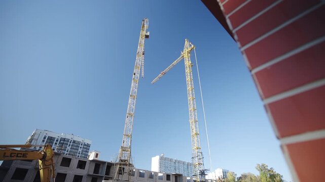 Powerful tower cranes assemble multistory apartment building under blue sky. Constructing machines work and lift cargoes in urban area