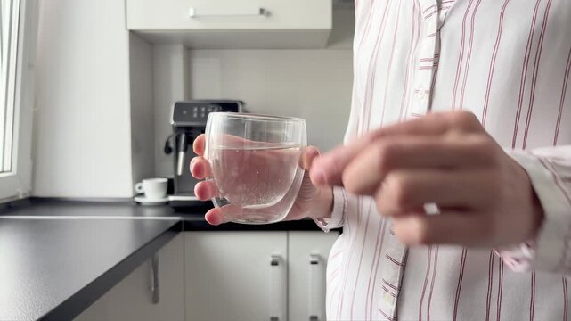 An unrecognizable young woman dissolves fizzy vitamins in a glass of water at home in the kitchen