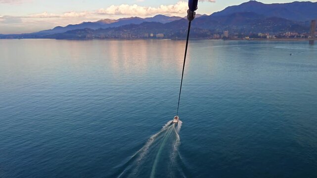 Aerial view of a speedboat pulling a parasail over calm sea, leaving wake trails. Scenic coastline with mountains and city in soft evening light, travel and adventure concept.