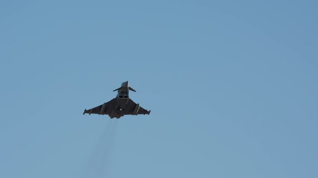 A military jet fighter flies overhead against a clear blue sky. The camera looks up from below, tracking the aircraft as it approaches