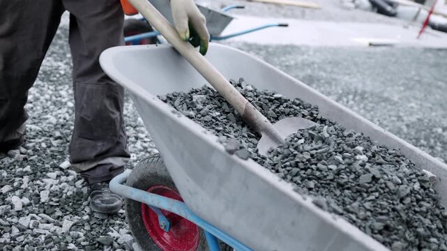 Builder in uniform takes gravel from wheelbarrow at construction site closeup. Laborer uses stone building materials outdoors slow motion