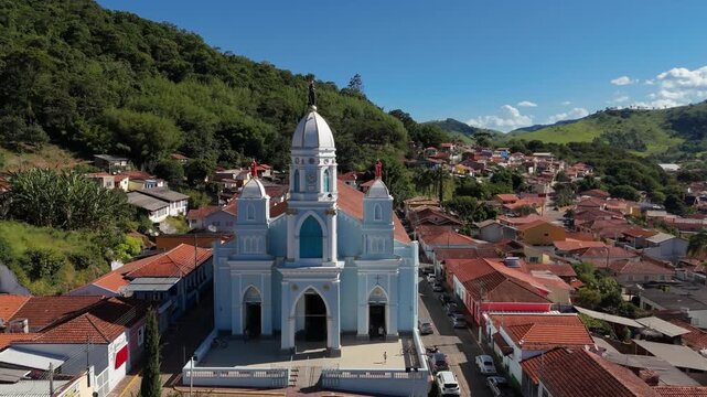 Aerial view of blue and white Matriz Church with belfries and dome in town center surrounded by red roof houses and green hills in Sao Bento do Sapucai, Brazil. Approach and crane down camera movement