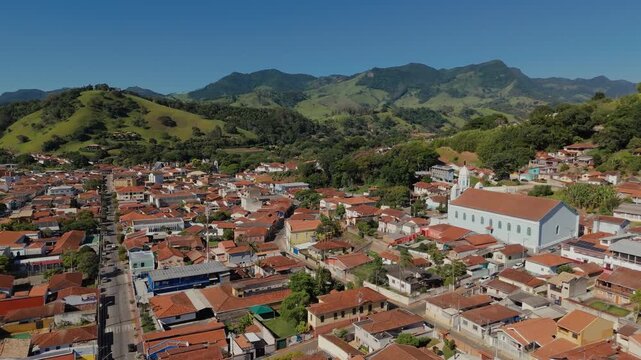 Aerial view of Matriz Church with dome and towers over residential area with red tile roofs and green mountains in background in Sao Bento do Sapucai, Brazil. Approach and overfly camera movement