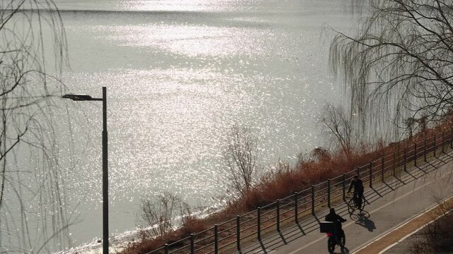 People Riding Bicycles on the Han River Path with Sparkling Sun Reflection on Water in Seoul