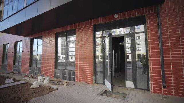 Open glass doors of incomplete apartment building at construction site. Stylish facade with brick decor and large windows in city