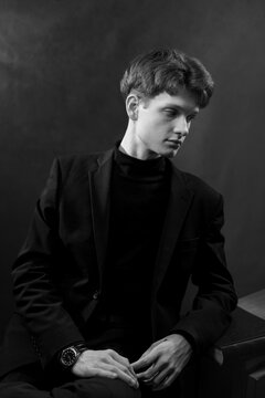 A cinematic black and white side-profile portrait of a young man with wavy hair. He is dressed in a sophisticated black blazer and turtleneck, sitting in a thoughtful pose against a dark background.