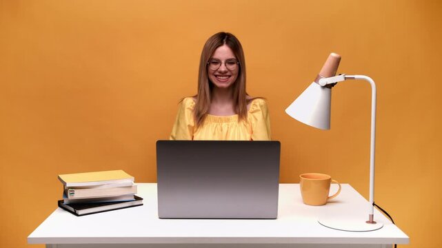 Smiling young professional woman with glasses typing on a laptop computer at a white desk against a vibrant yellow background