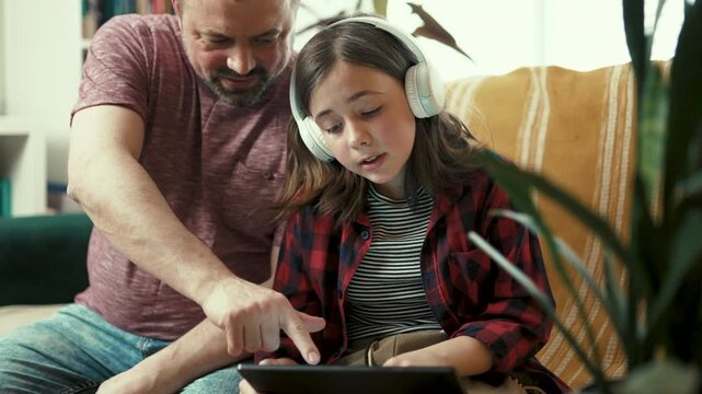 Preteen girl in headphones showing interesting content on digital tablet to her father at home-4k,10 bit footage.