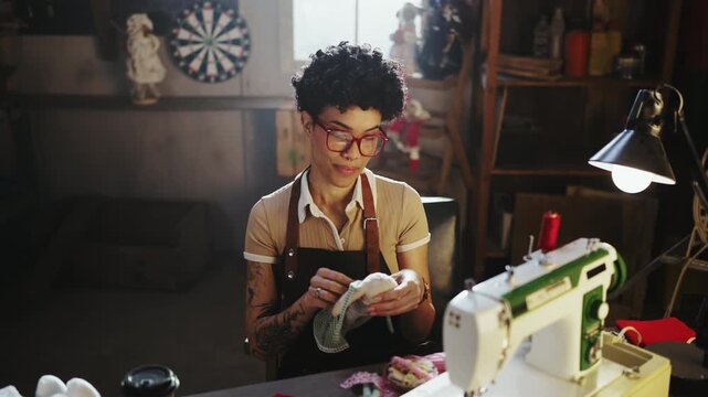 A person is sewing fabric in a workshop. The room has shelves and tools. The individual is focused on their task and handling materials at a sewing machine.