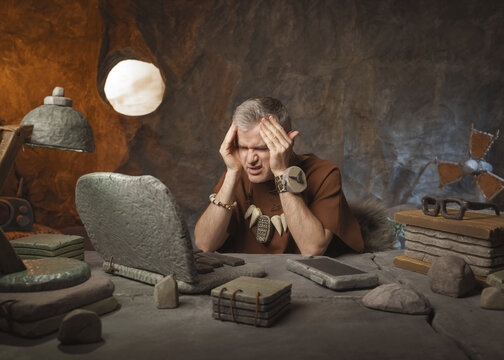 Prehistoric Burnout: Funny Caveman with Headache Working on Stone Laptop in Cave Office