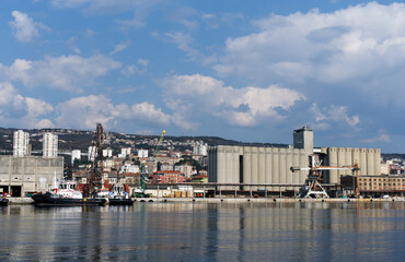 Obraz premium Industrial landscape of a commercial sea port featuring large concrete grain elevator silos for agricultural export. The scene includes cargo terminal infrastructure, harbor cranes, moored tugboats