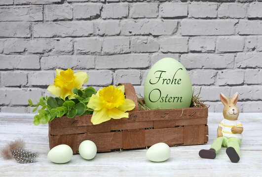 Spankorb mit gelben Narzissen, gr&uuml;nem Blattwerk und einem gro&szlig;en Ei mit der Aufschrift 'Frohe Ostern' daneben ein sitzender Osterhase und  kleine Eier auf hellem Untergrund vor wei&szlig;er Ziegelwand.